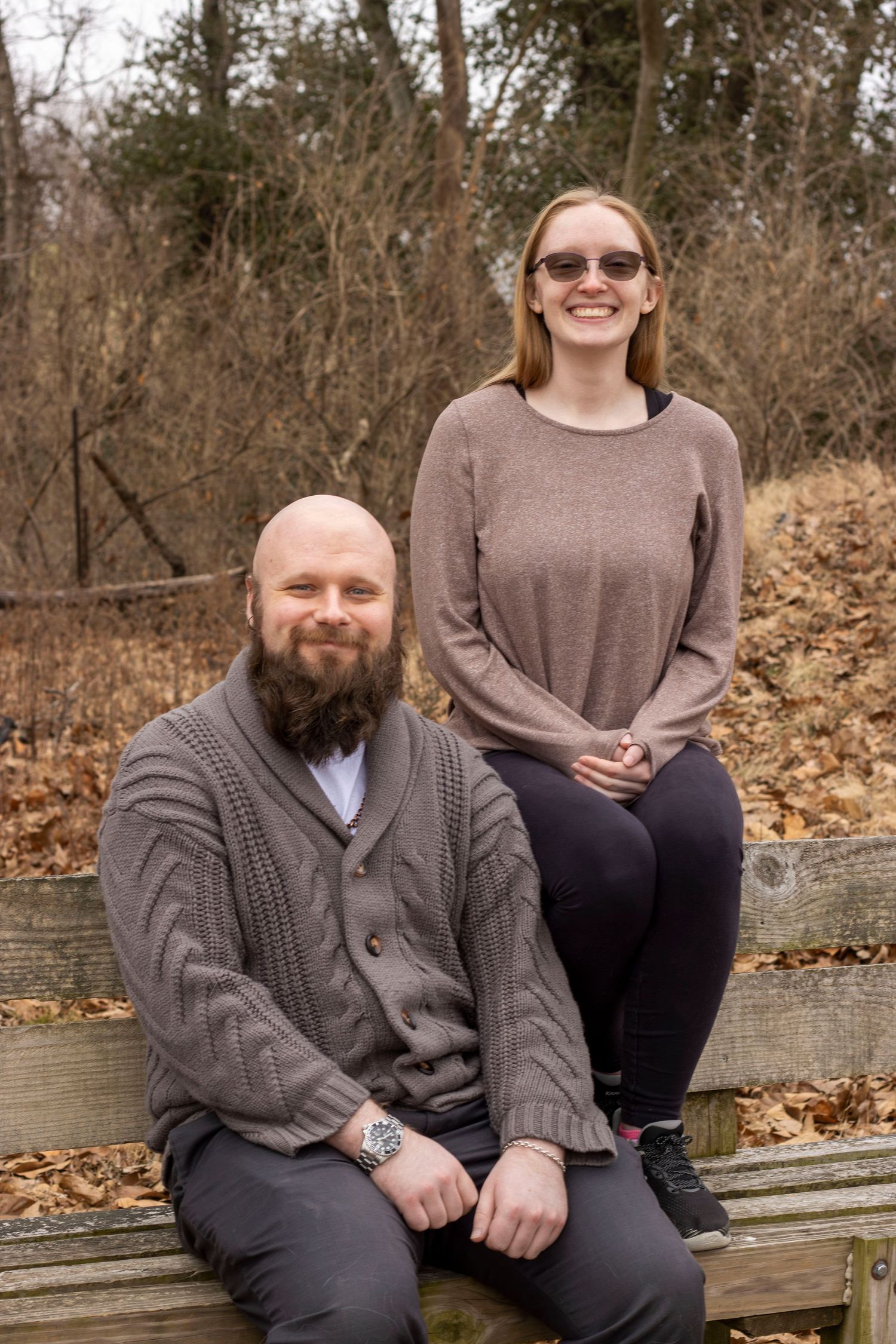 Two people sitting on a wooden bench in autumn with fallen leaves on the ground in a natural outdoor setting.