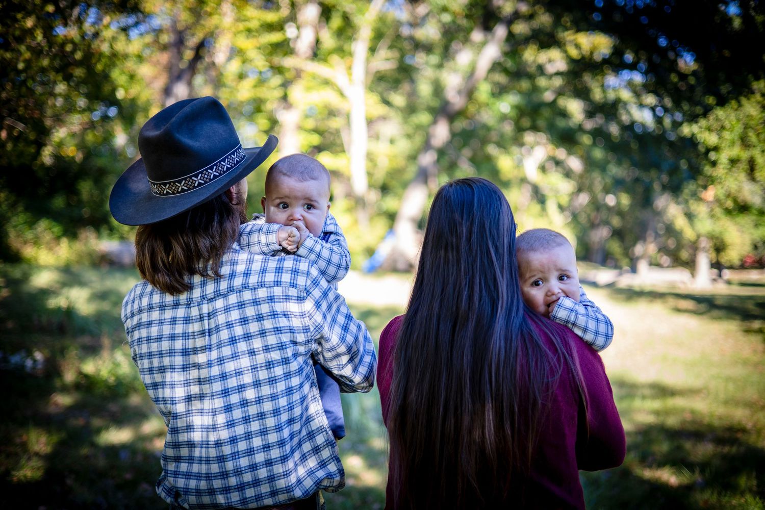 Two parents seen from behind hold their babies outdoors in a sunlit park, one wearing a plaid shirt and hat, the other with long dark hair.