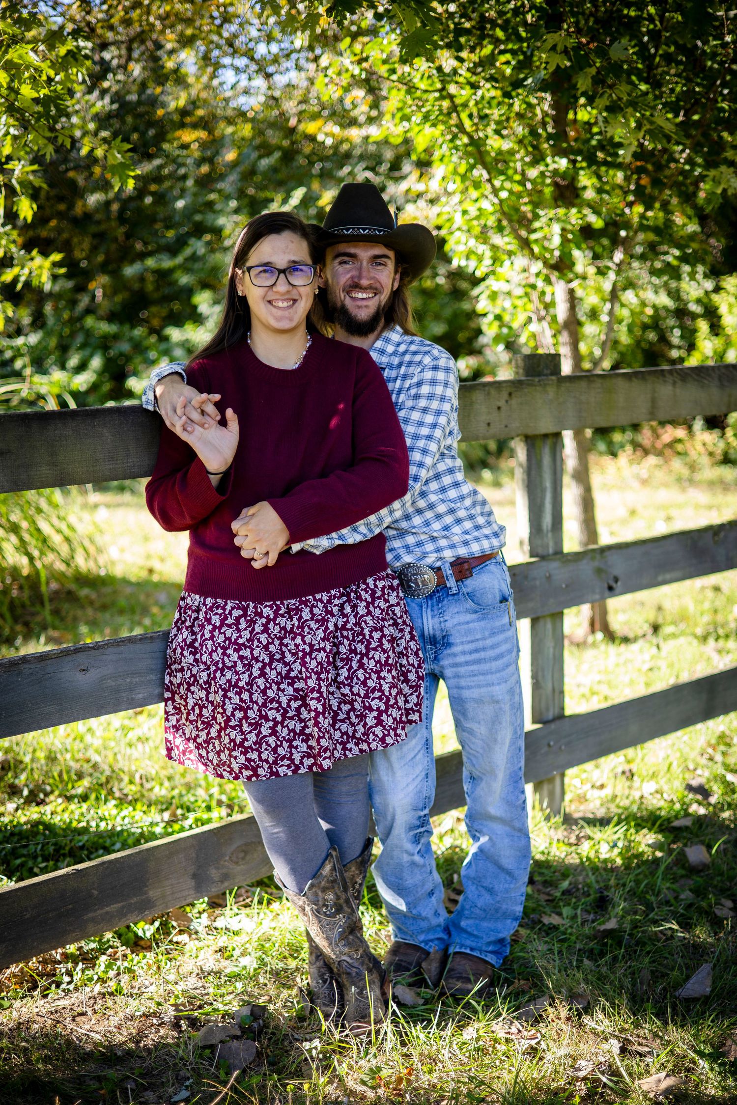 Smiling couple in western clothing and cowboy hat embrace beside a rustic wooden fence surrounded by green trees on a sunny day.