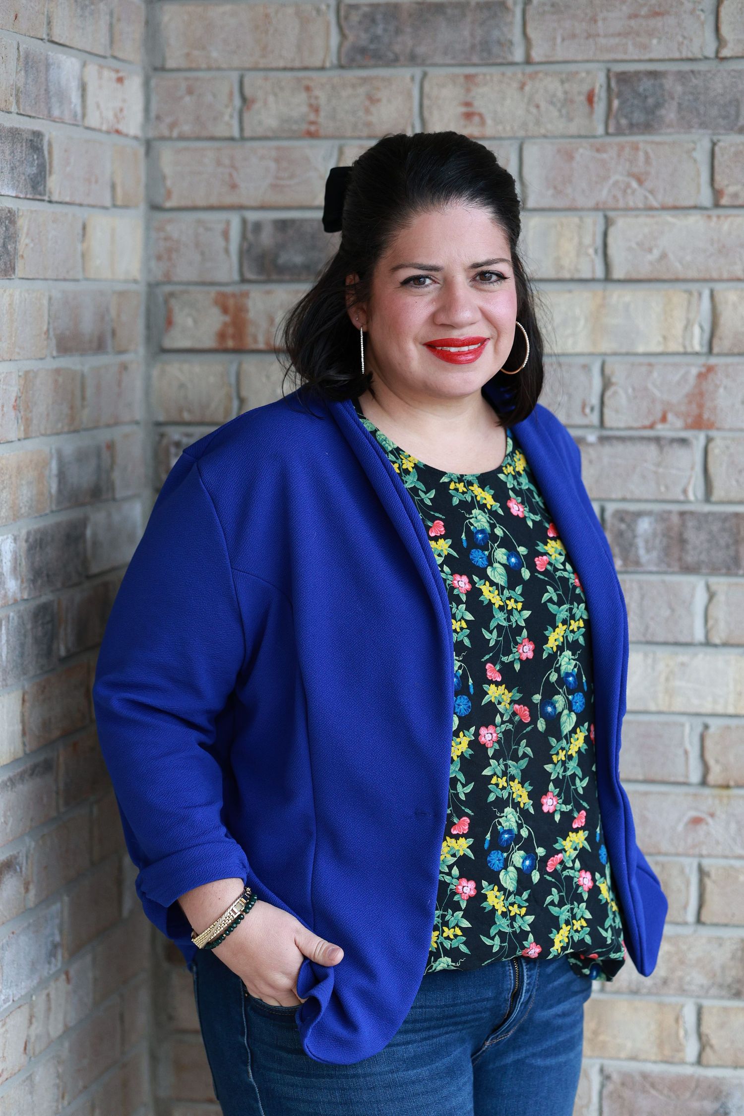 Woman in blue blazer and floral top smiling against brick wall, wearing hoop earrings and red lipstick.