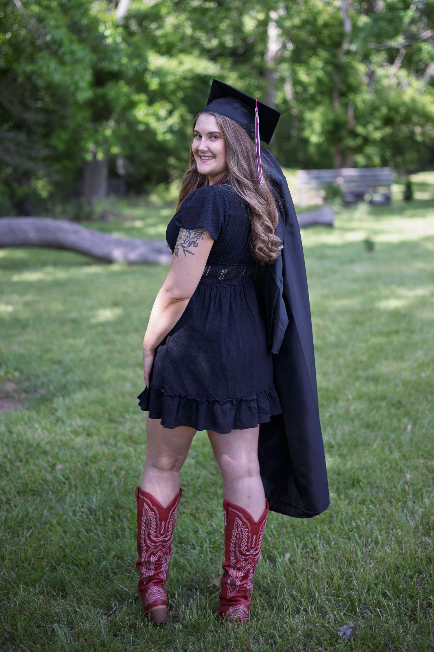 Graduate in black cap and gown wearing red cowboy boots and black dress, posing outdoors on green grass.