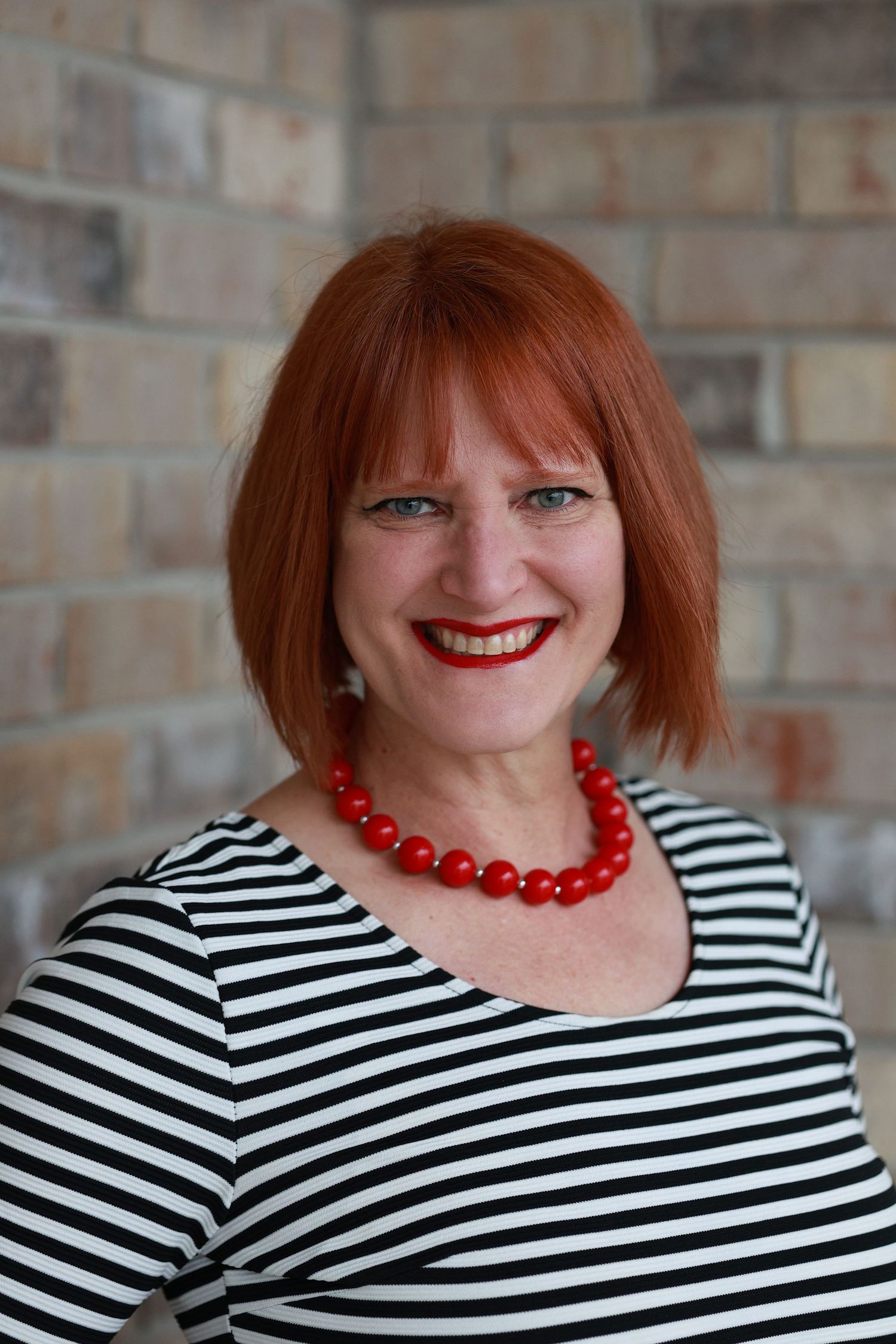 A smiling woman with red hair wearing a black and white striped top and bold red beaded necklace against a brick wall.