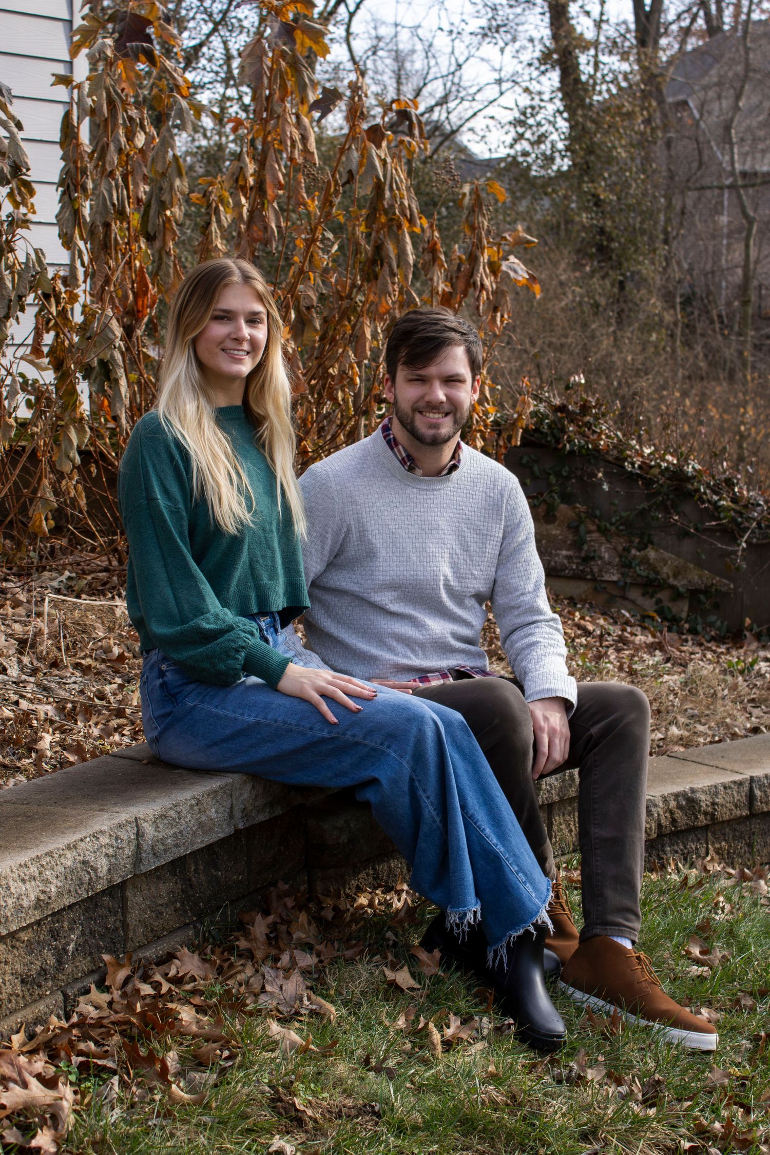 A couple sits together on a stone wall on an autumn day with dried leaves and foliage in the background.