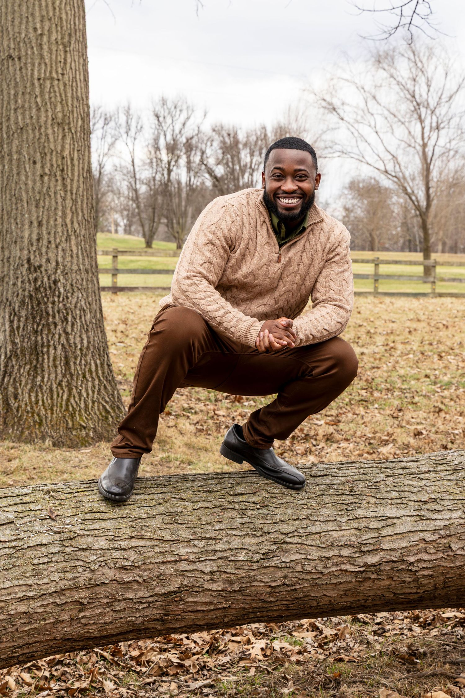 Person in tan sweater sits casually on fallen tree trunk in autumn park setting with brown leaves on ground.