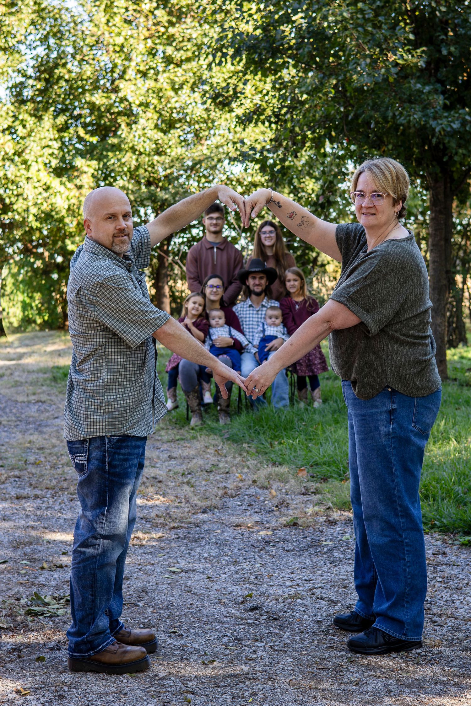 A man and woman form a heart shape with their arms in the foreground while a family poses together on a tree-lined gravel path.
