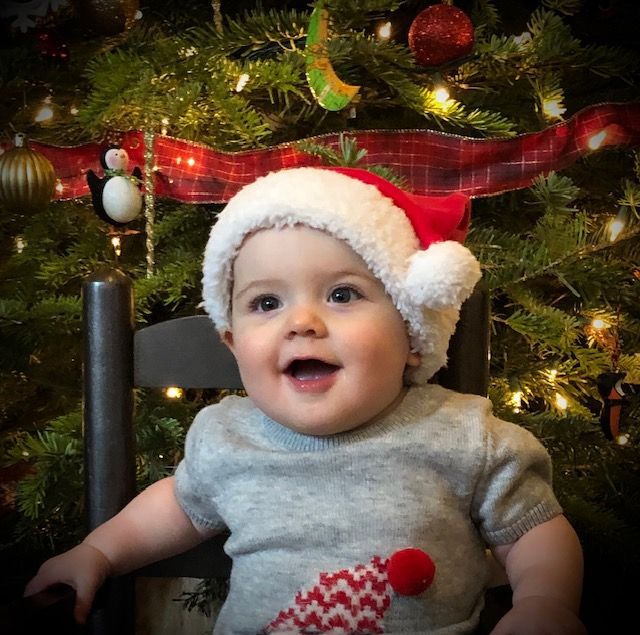 A small child wearing a Santa hat and gray shirt smiles joyfully in front of a decorated Christmas tree.