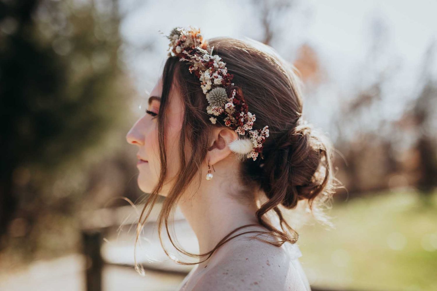 A bride in a lace wedding dress poses outdoors in soft natural lighting with floral hair accessories.