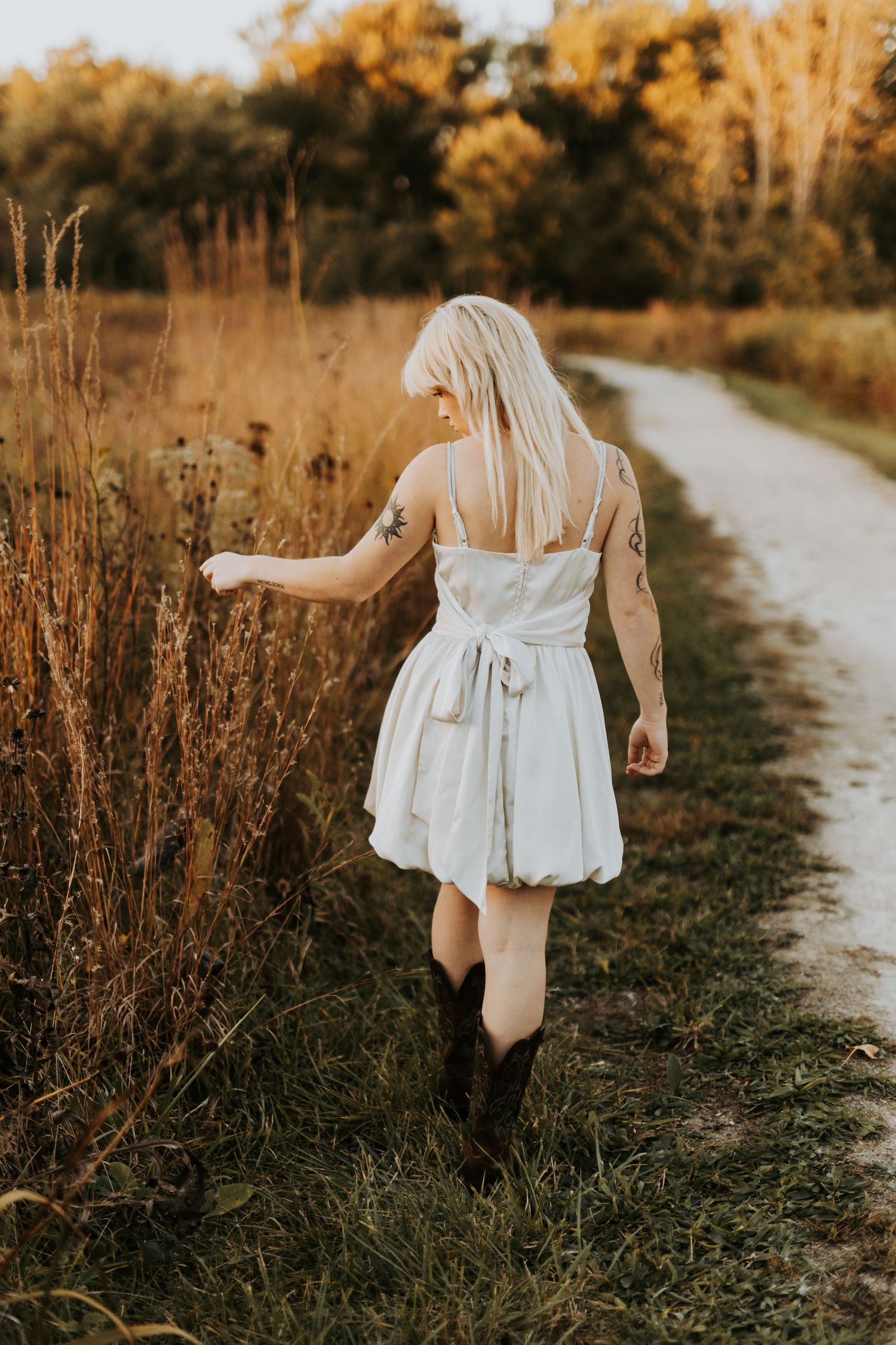 A person in a white dress walks along a winding dirt path through golden autumn fields at sunset.