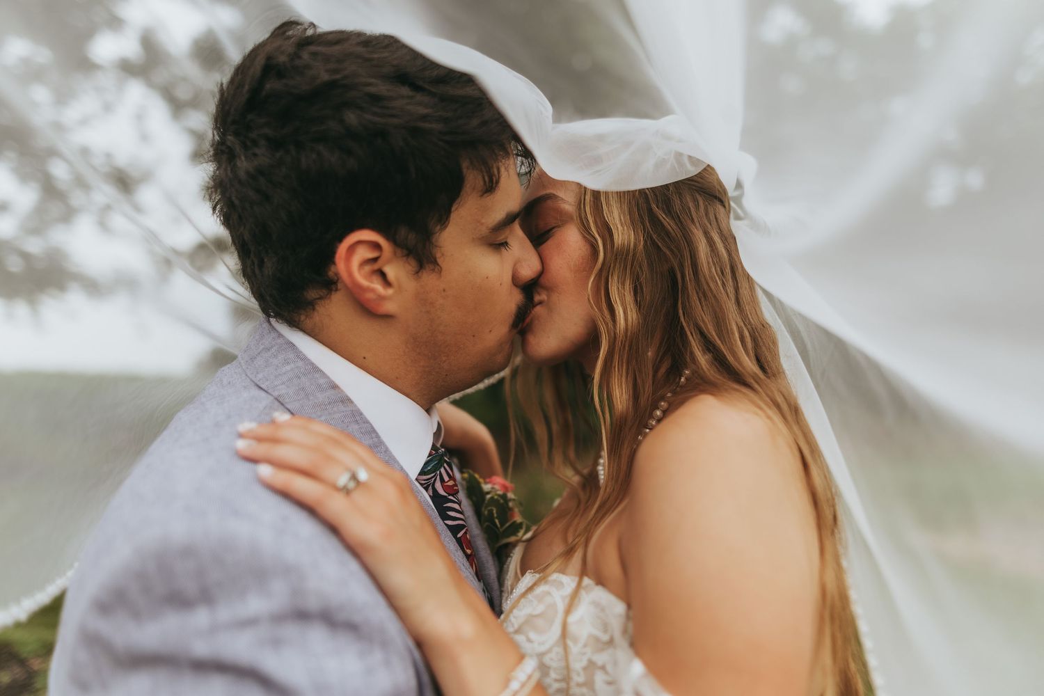 Bride and groom kissing under a flowing veil outdoors on their wedding day.
