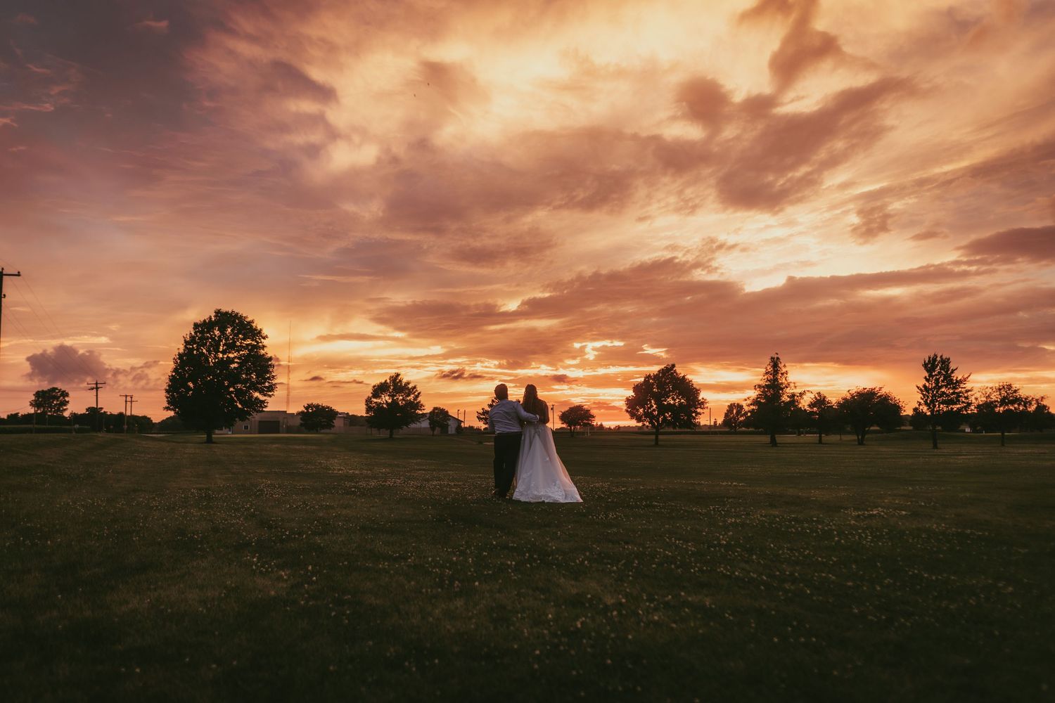 Bride and groom embracing at sunset on open green field with dramatic orange sky.