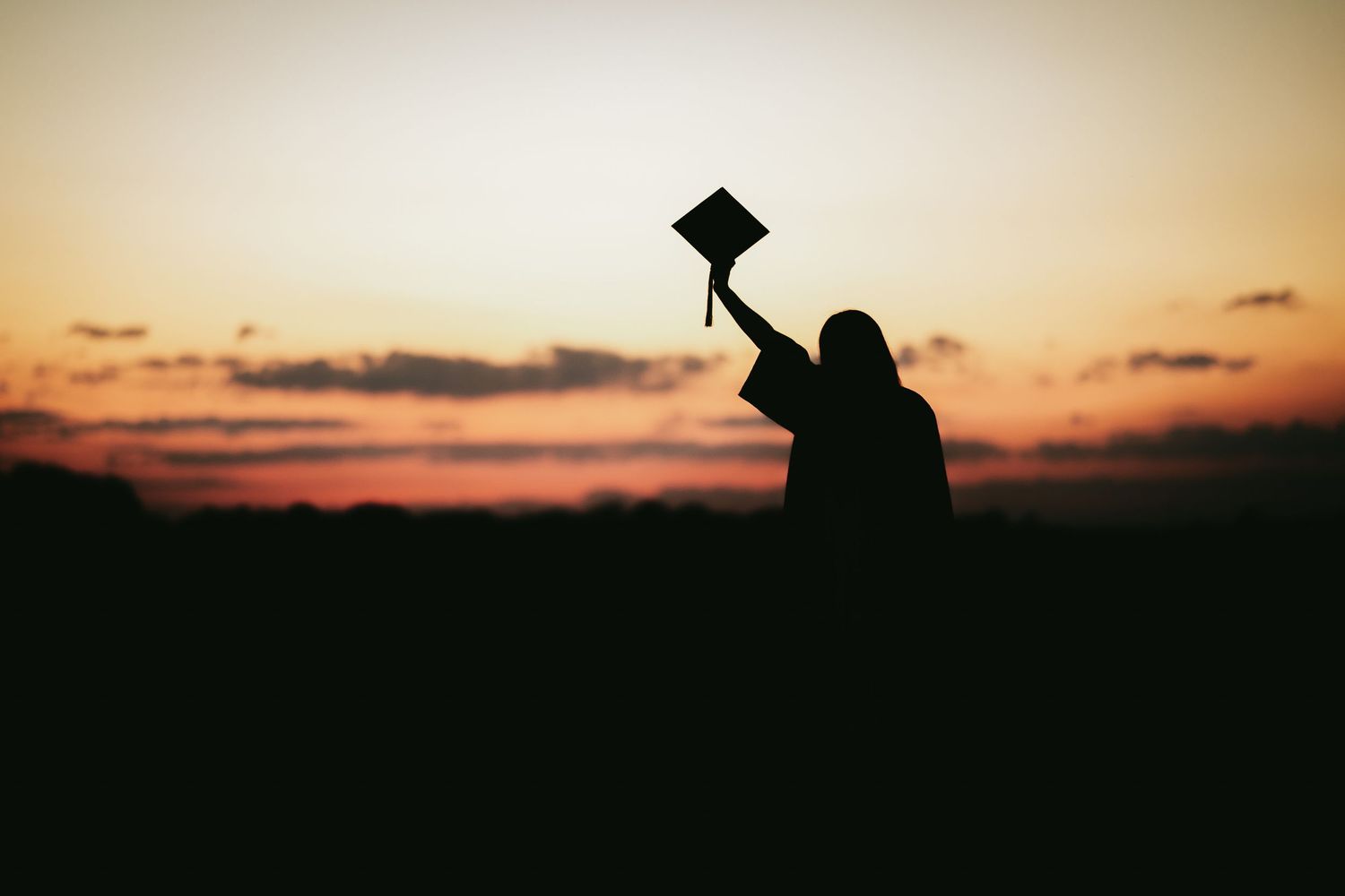 Silhouette of a graduate holding up diploma against vibrant orange sunset sky.