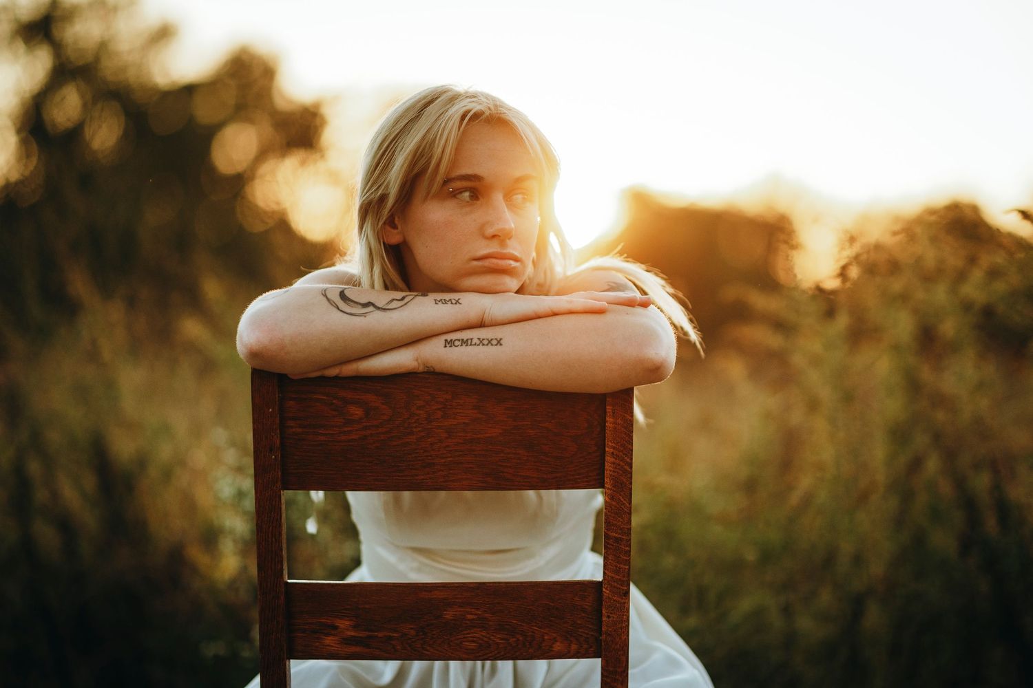 A person sits on a wooden chair in a golden sunlit field during sunset, creating a dreamy atmospheric scene.