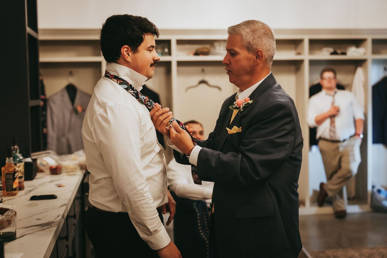 Father helping groom tie bow tie in dressing room before wedding ceremony.