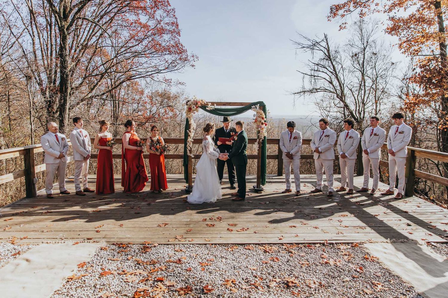 A wedding party stands in a line outdoors during autumn with fallen leaves scattered on the ground.
