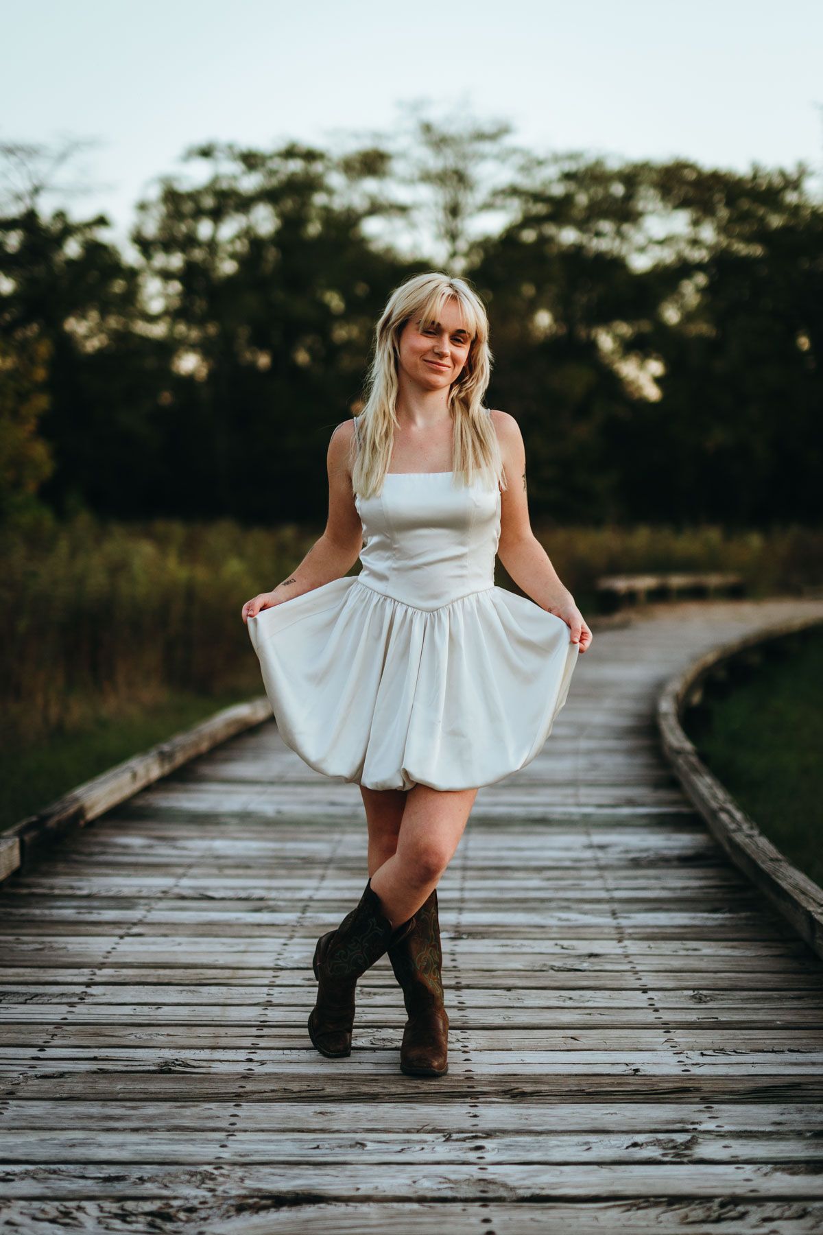 A person in a white dress poses playfully on a wooden boardwalk during golden hour.