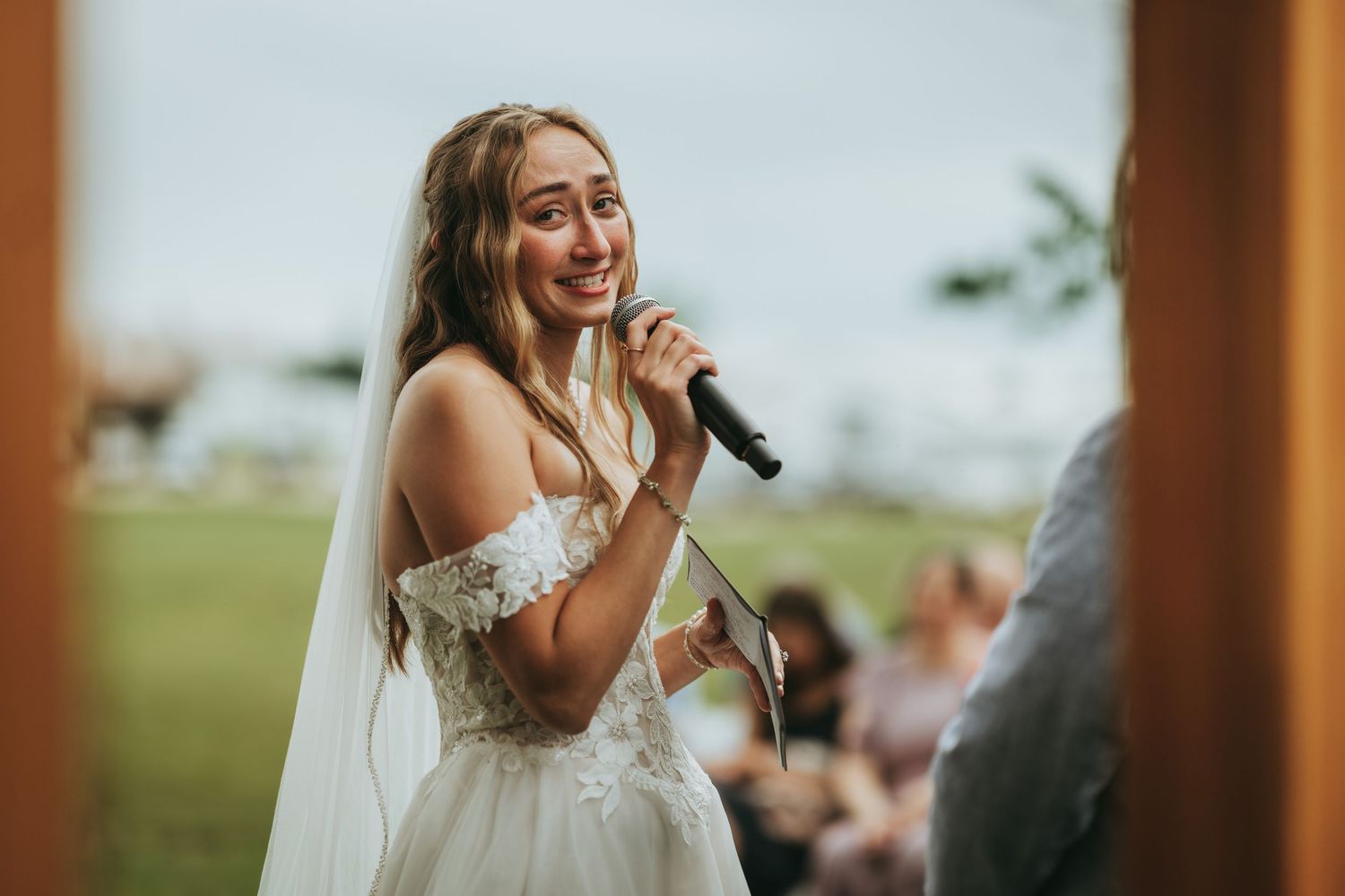 Smiling bride in lace off-shoulder gown speaking into microphone during outdoor wedding ceremony.