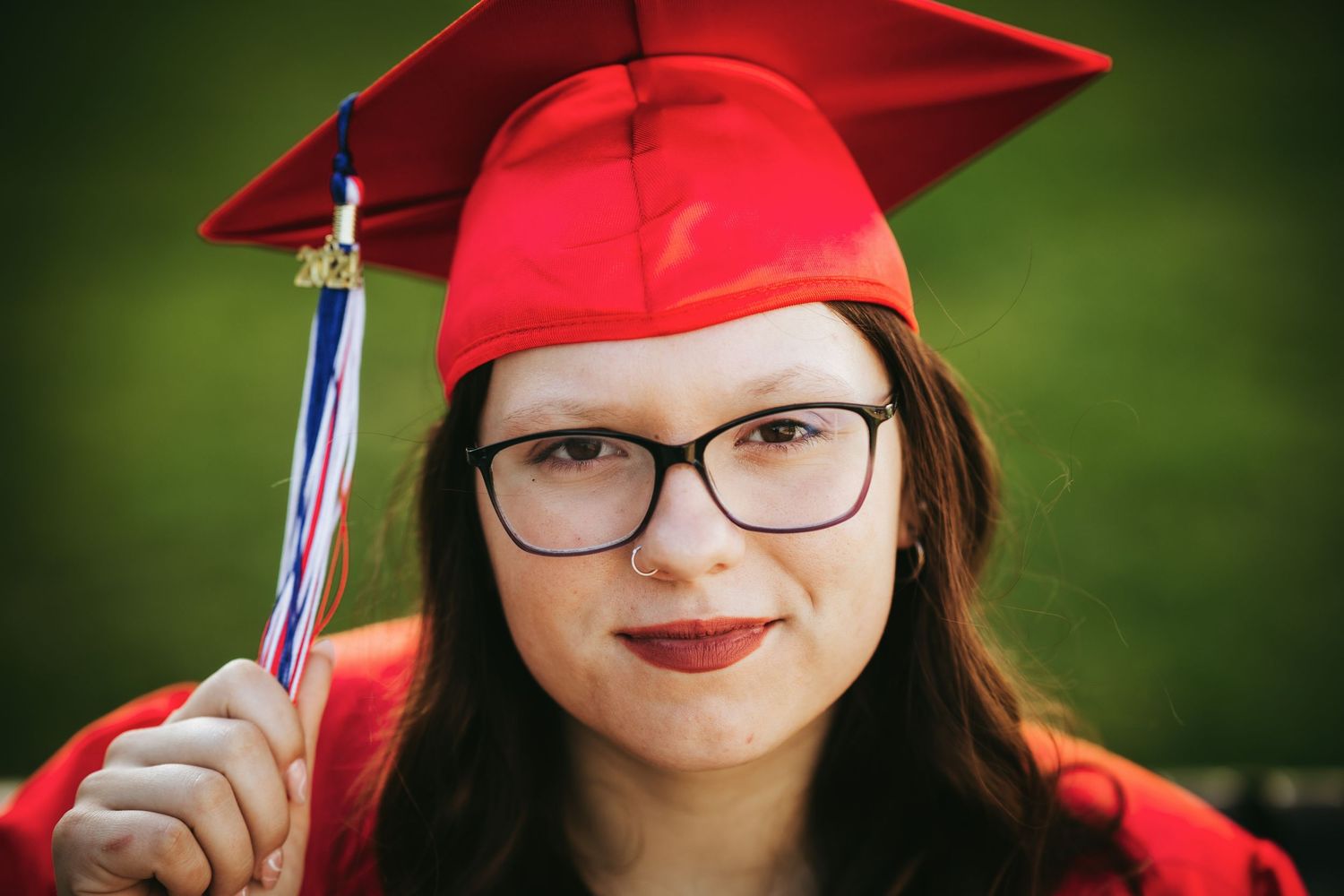 Graduate in red cap and gown holds diploma while smiling outdoors on graduation day.