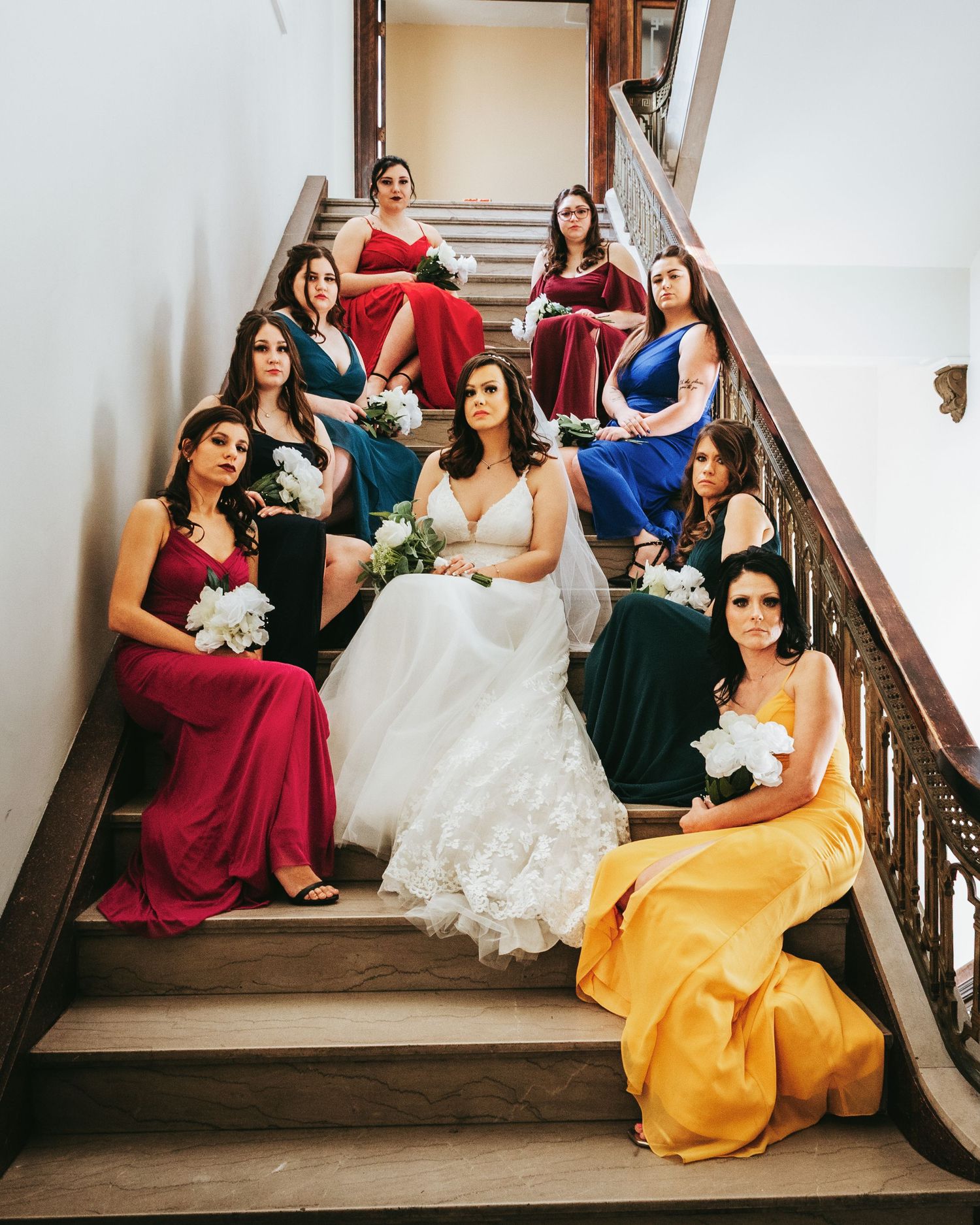 Bridesmaids in burgundy and yellow dresses pose elegantly on a grand staircase with the bride in a white gown.