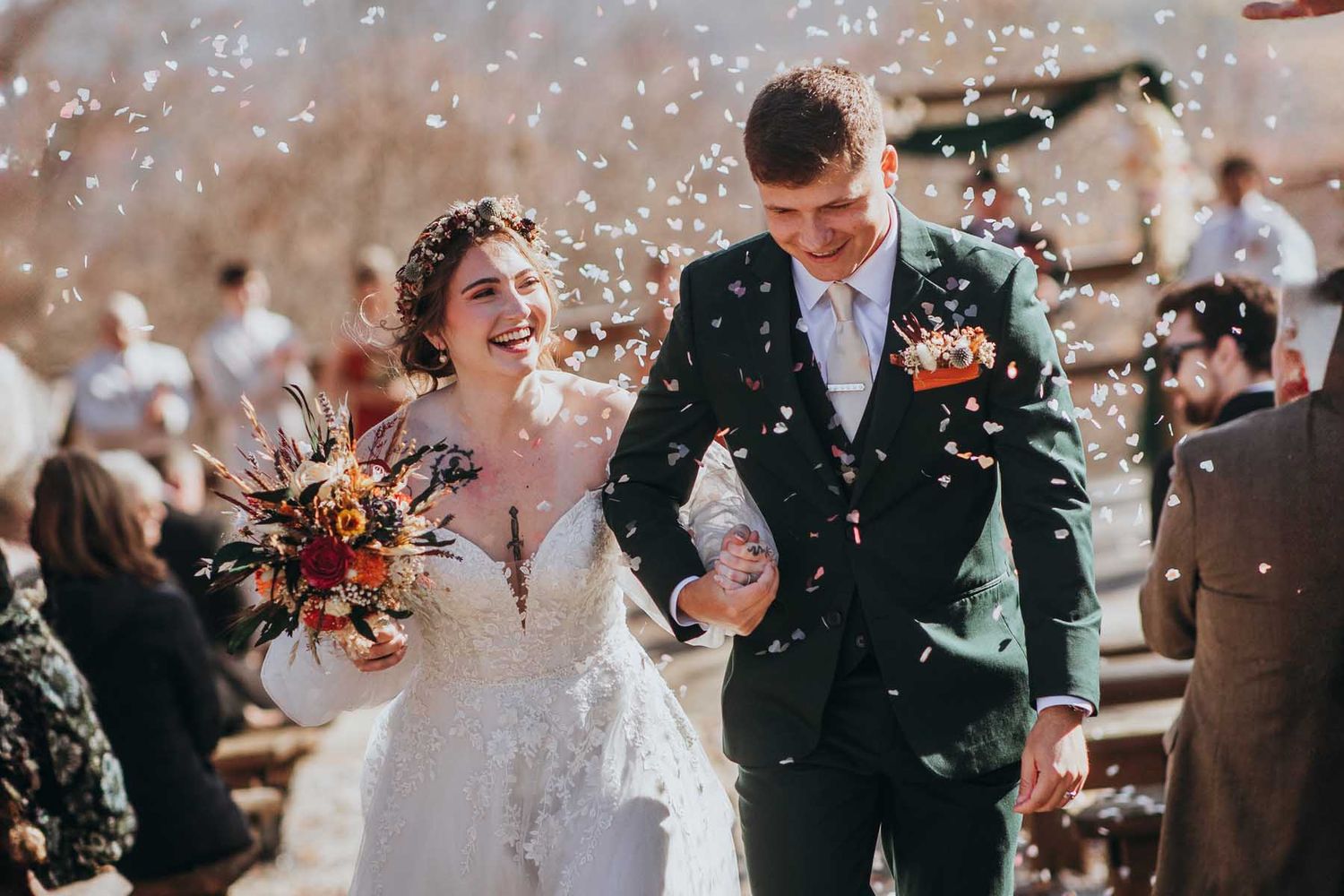 A joyful couple walks through a shower of confetti after their wedding ceremony while holding autumn-colored flowers.