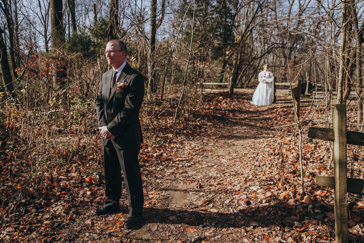 A couple in formal attire walks together through a wooded area covered in fallen autumn leaves.