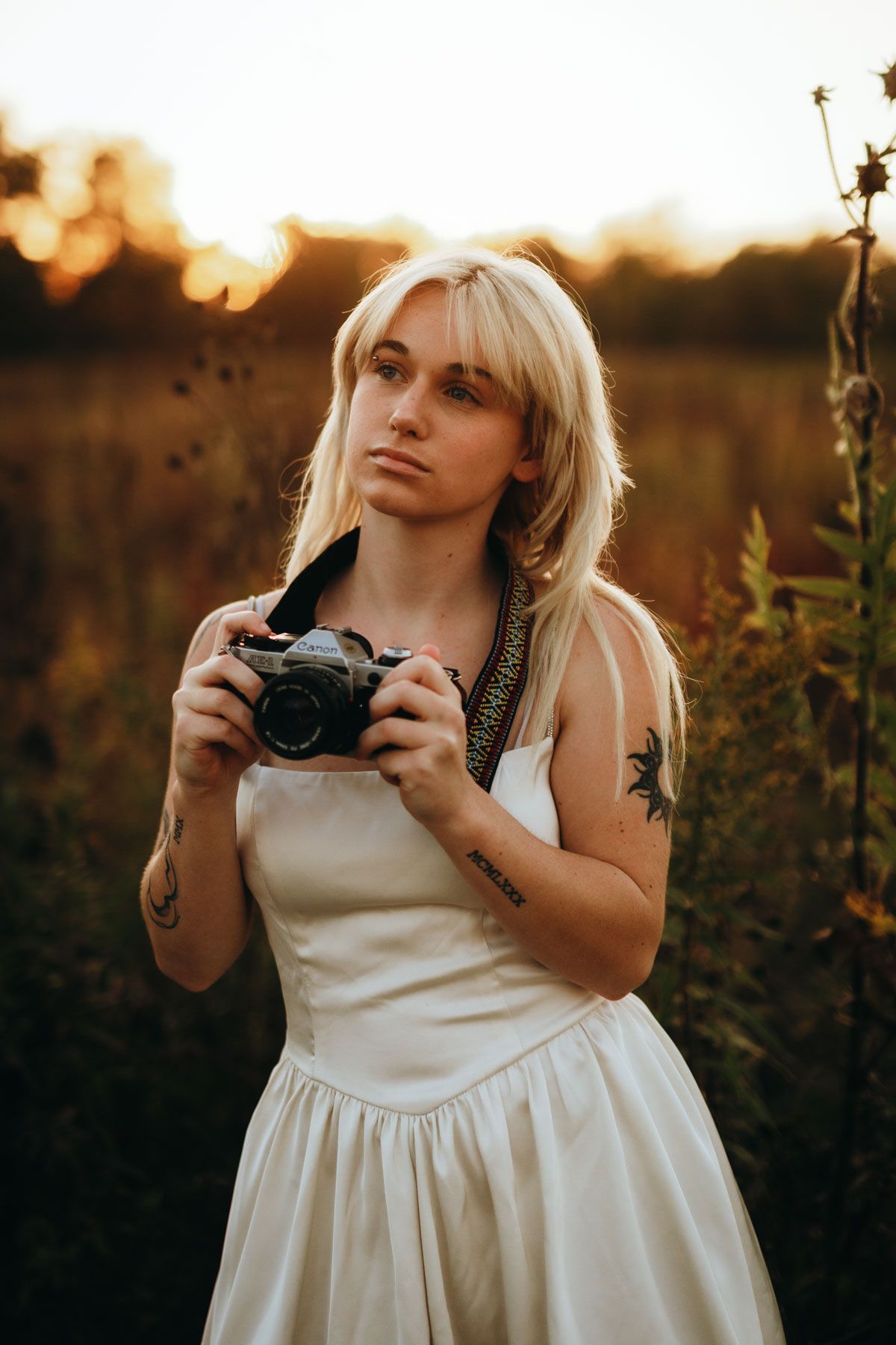 Photographer in white dress holding camera during golden hour sunset in field with wildflowers.