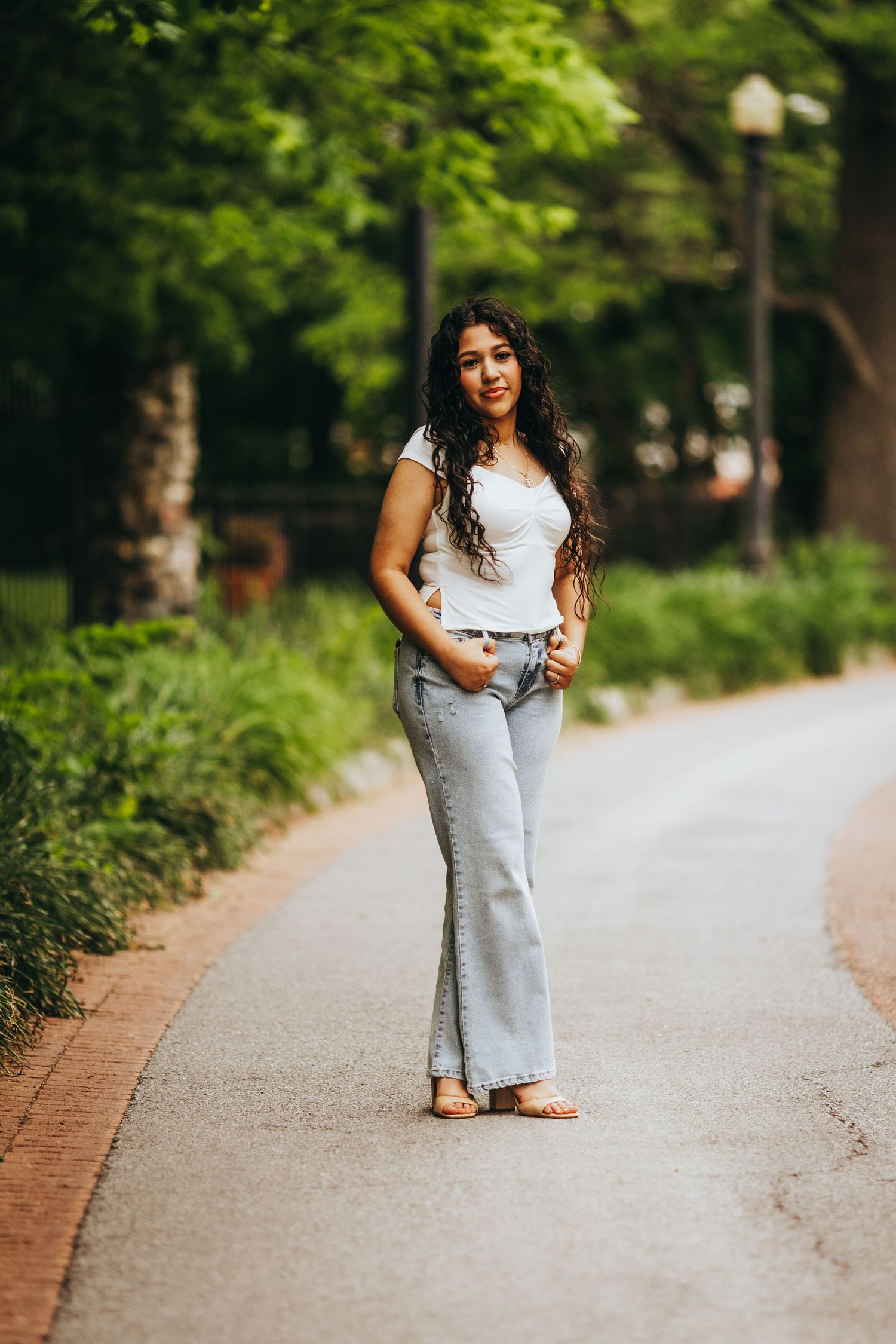 A person wearing a white top and light denim jeans poses casually on a tree-lined path in a park setting.