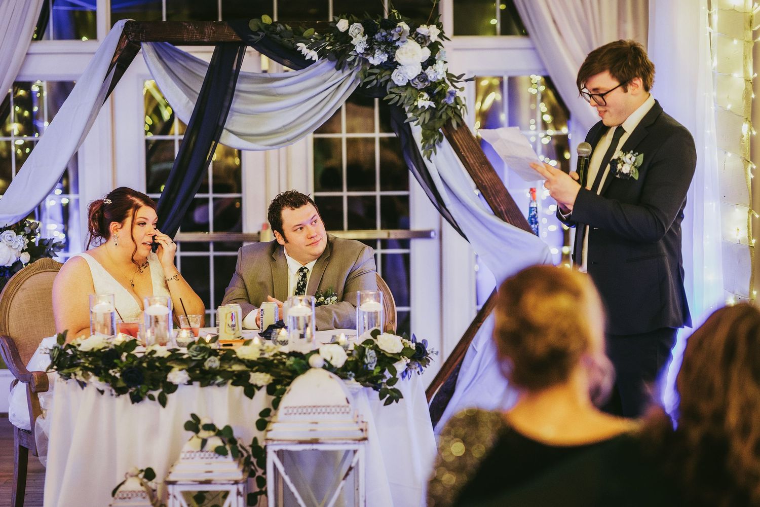 A wedding toast being given at an indoor reception with elegant white and floral decorations.