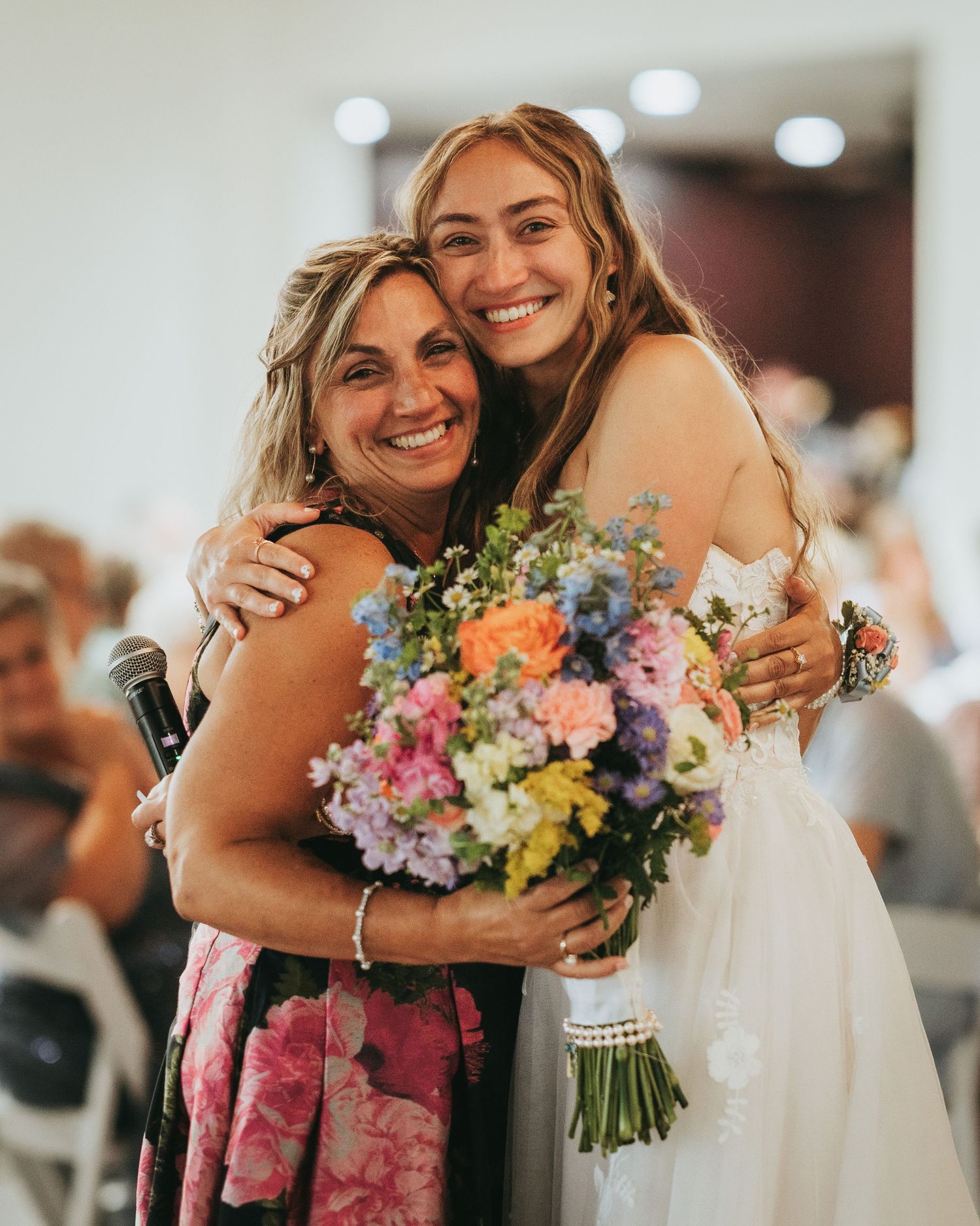 Bride in white gown hugs smiling woman holding colorful bouquet at wedding reception.