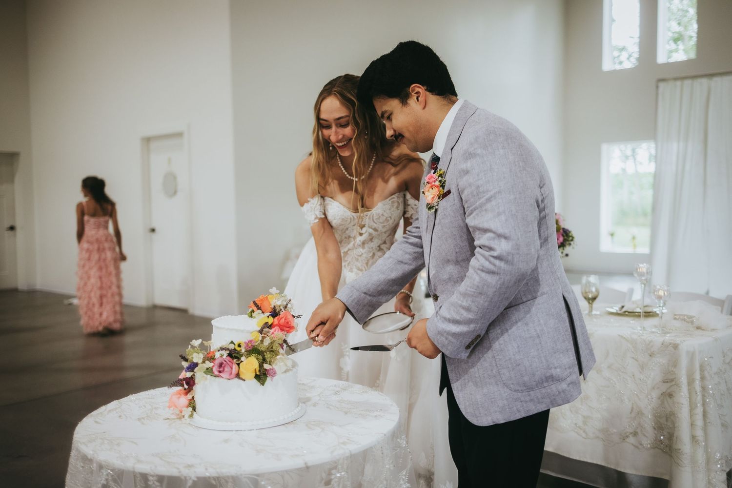 Newlyweds slice their floral-adorned white wedding cake together at bright indoor reception venue.