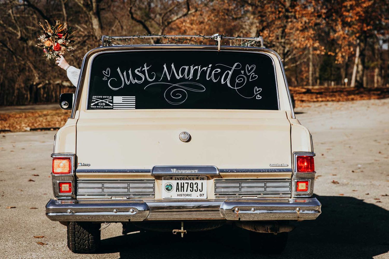 A vintage car with 'Just Married' text written on its rear window drives away on an autumn road with fallen leaves.