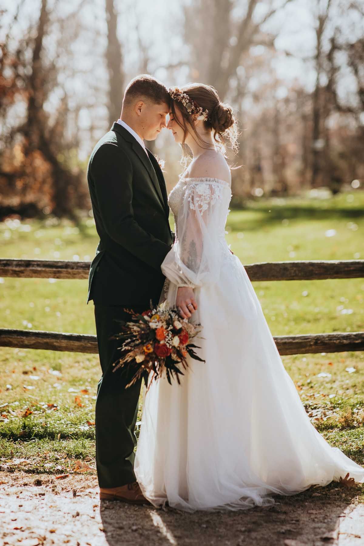 A romantic wedding couple stands together in autumn sunlight by a rustic wooden fence holding a fall-colored bouquet.