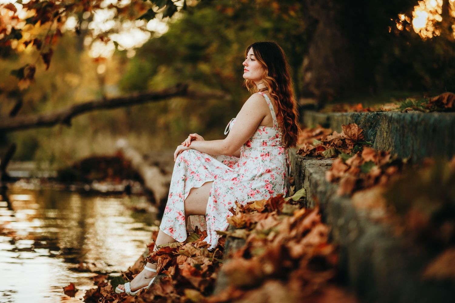 A woman in a floral dress sits peacefully by a pond surrounded by autumn leaves at sunset.