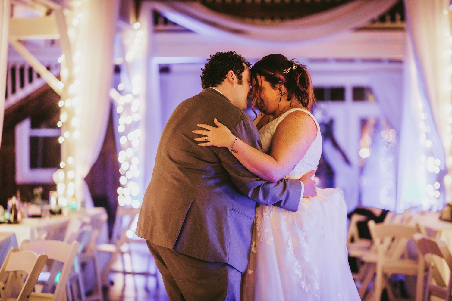 Couples dancing at wedding reception under purple ambient lighting and white draped decorations.