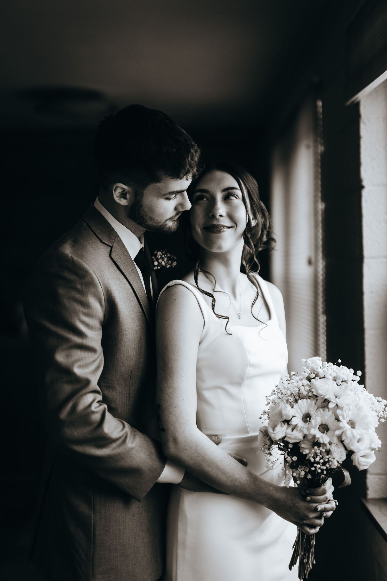 Black and white wedding photo showing couple in formal attire sharing an intimate moment while holding a floral bouquet.
