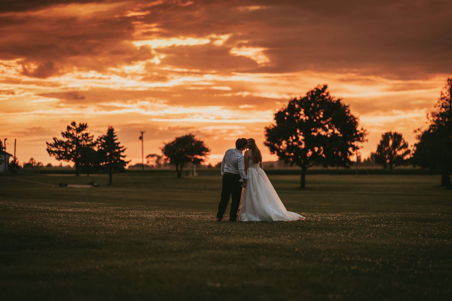 Bride and groom share romantic kiss at sunset on green lawn with dramatic orange sky.