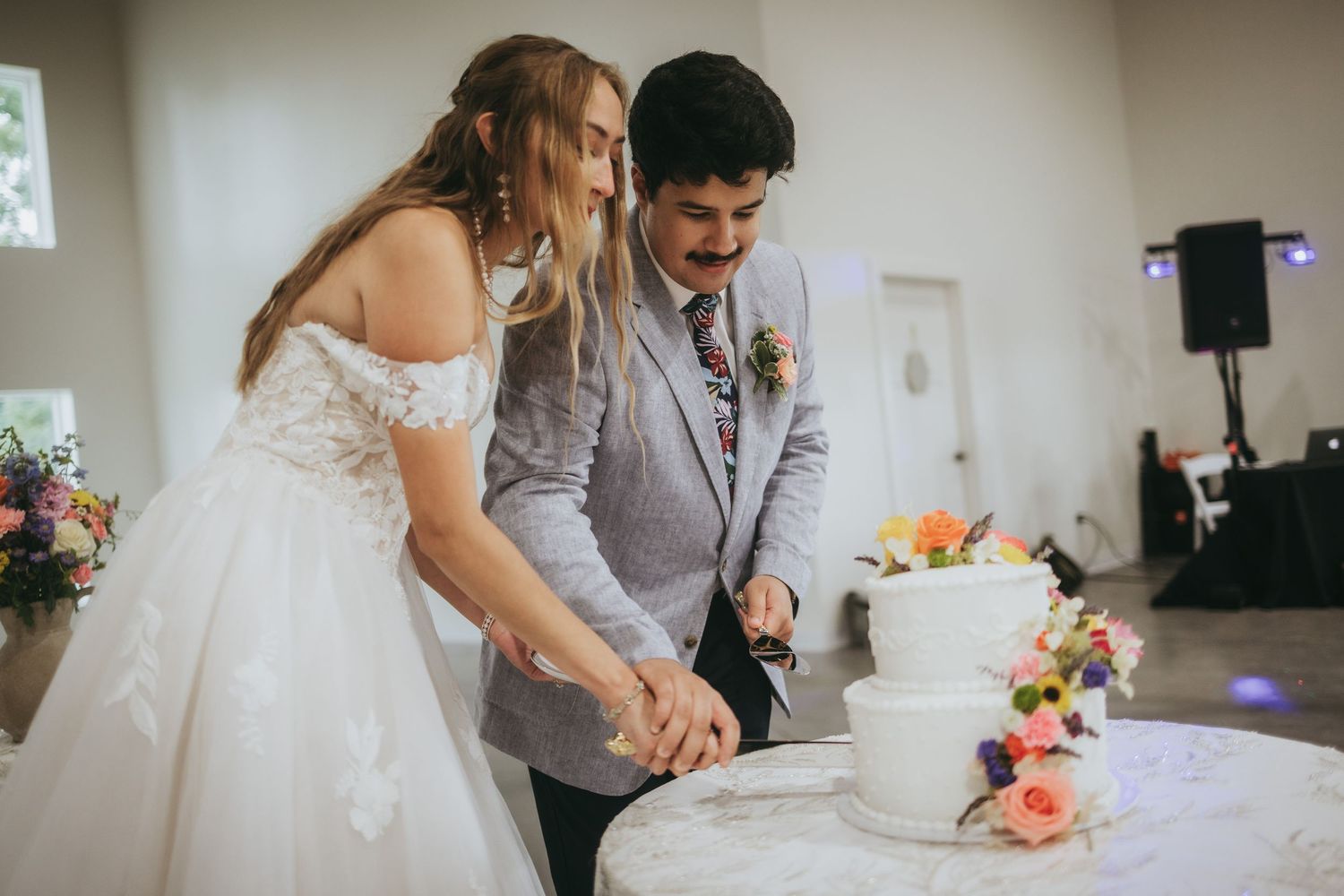 Bride and groom cut elegant white wedding cake decorated with colorful flowers at reception.