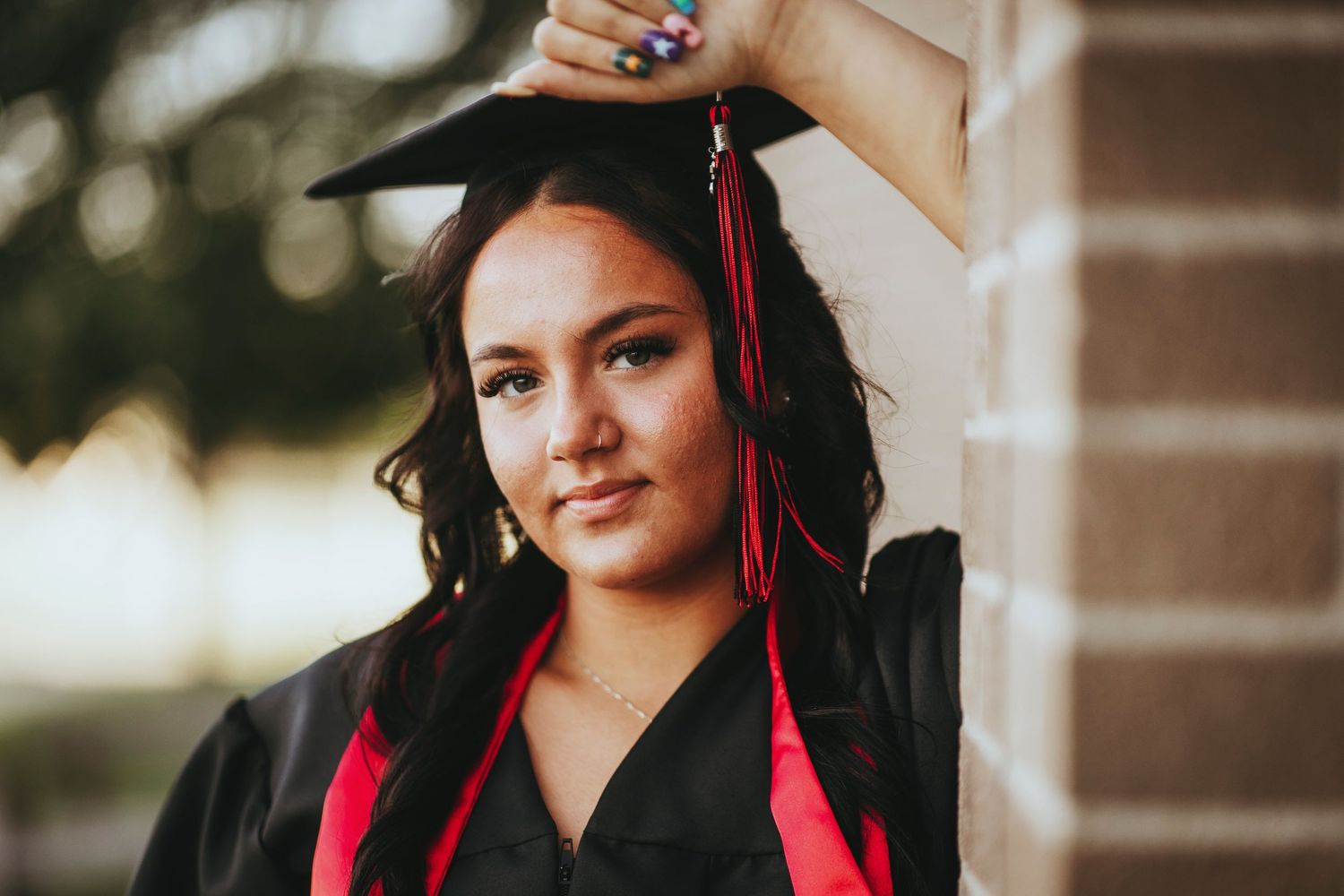 Graduate in black cap and gown with red tassel holds graduation cap against blurred outdoor background.