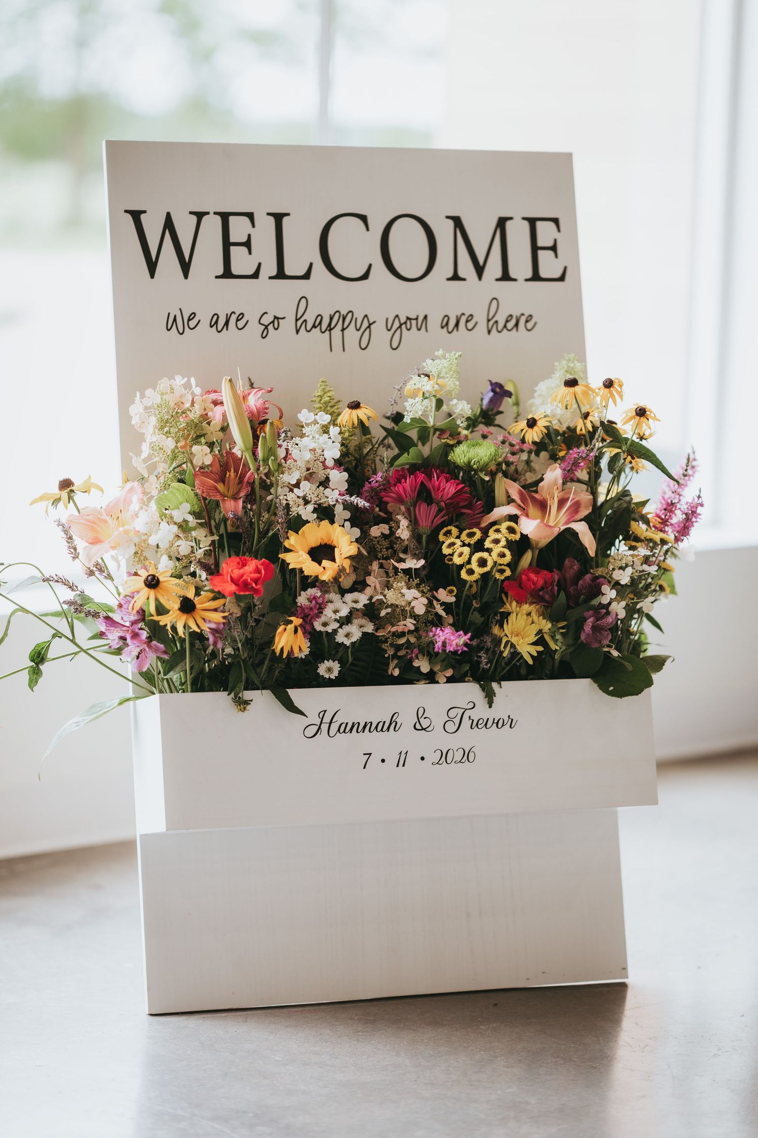 Wedding welcome sign with wildflowers for Hannah & Trevor, dated 7-11-2026, reading we are so happy you are here.