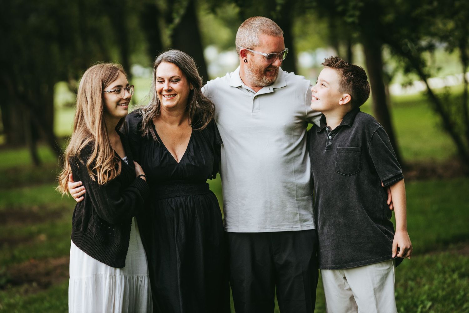 A happy family of four laughs together while posing outdoors in a park setting with green trees in the background.