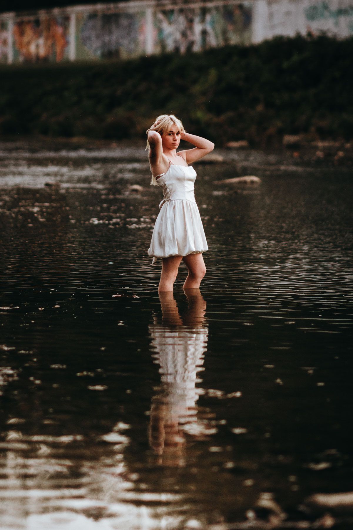 A figure in a white dress wades through dark reflective water in a moody nature setting.