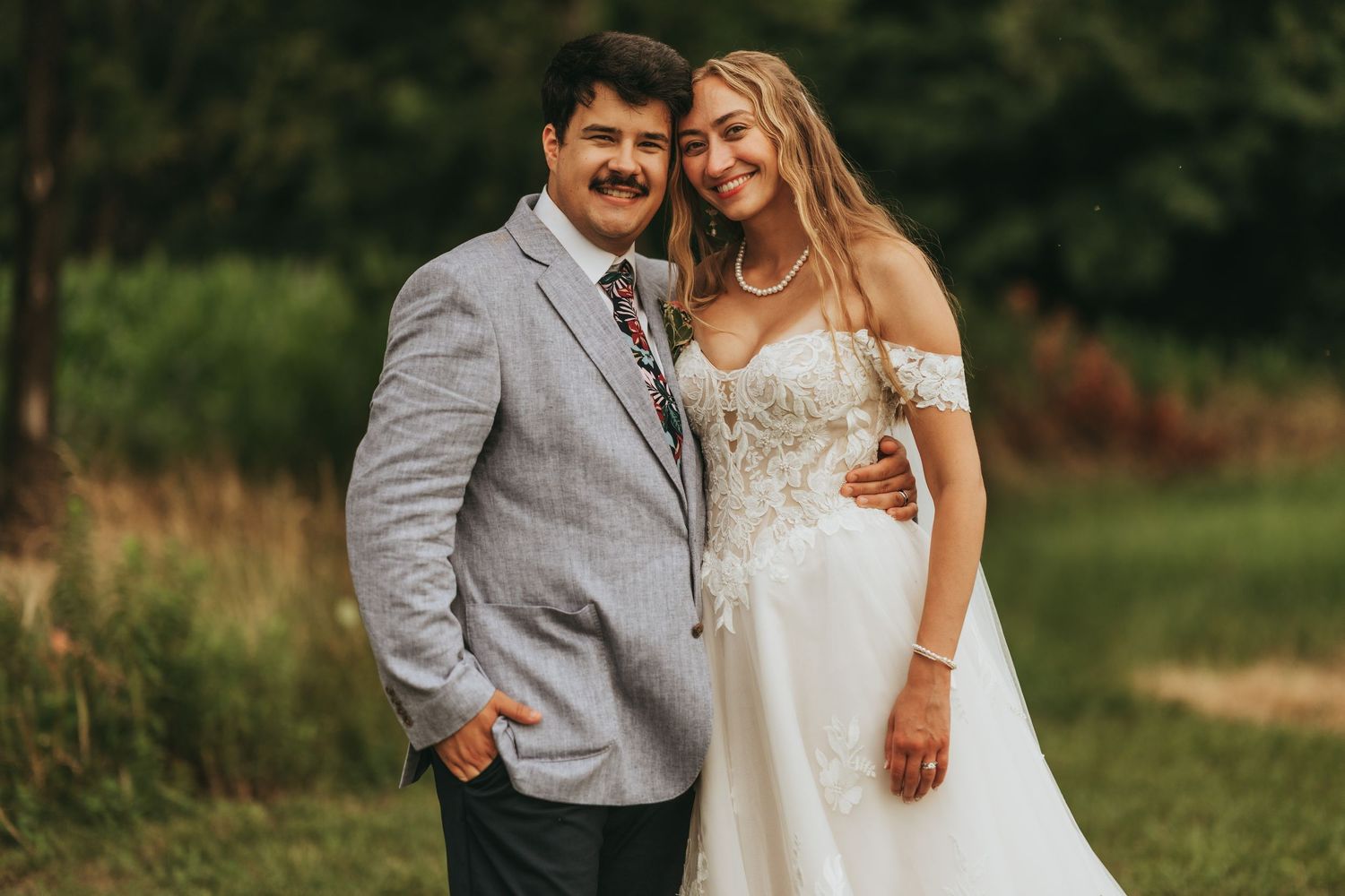 A bride in an off-shoulder white gown poses with a man in a gray suit outdoors at a wedding celebration.