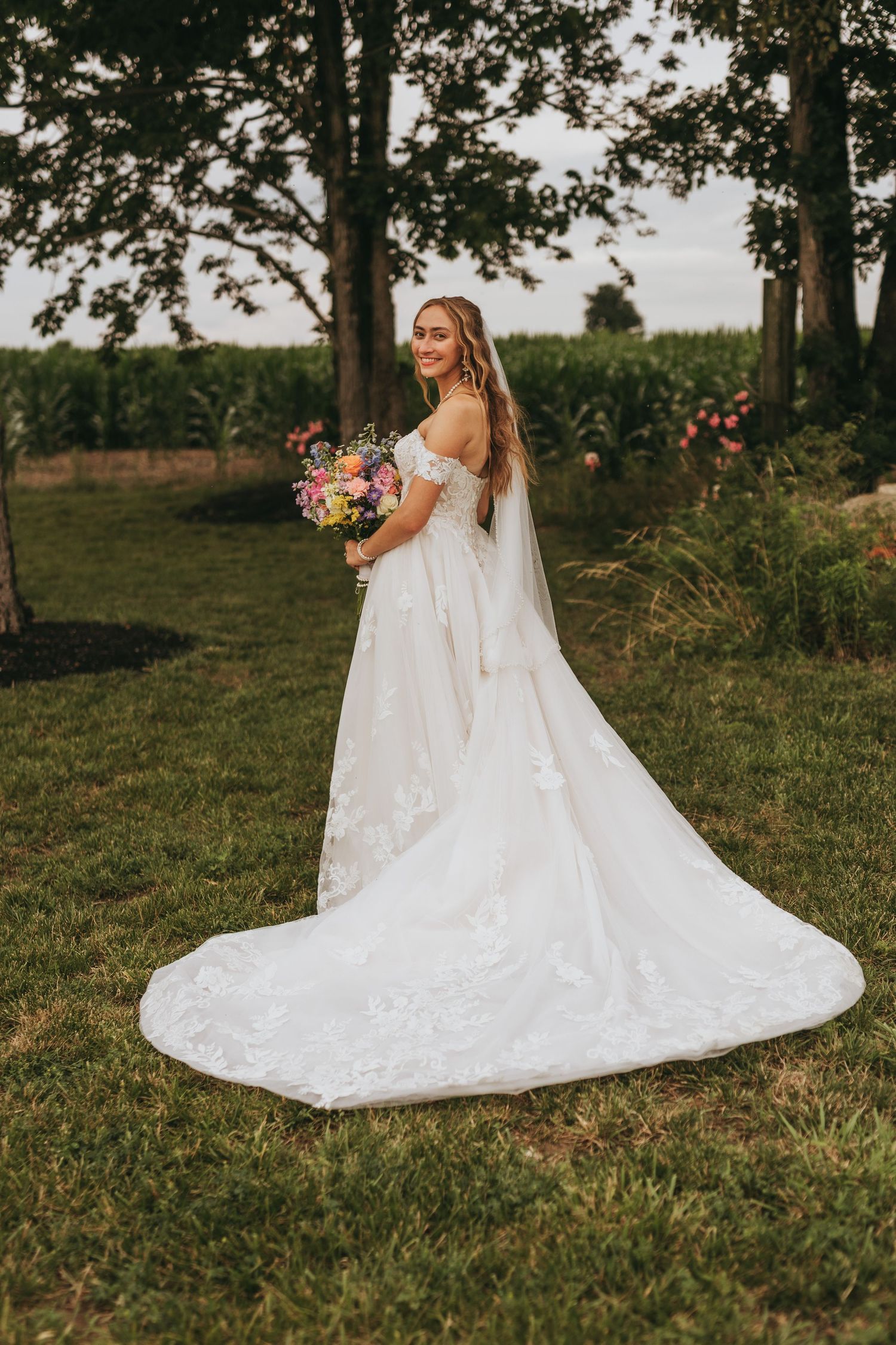 Bride in white off-shoulder lace gown with train holds colorful bouquet outdoors near trees and cornfield.