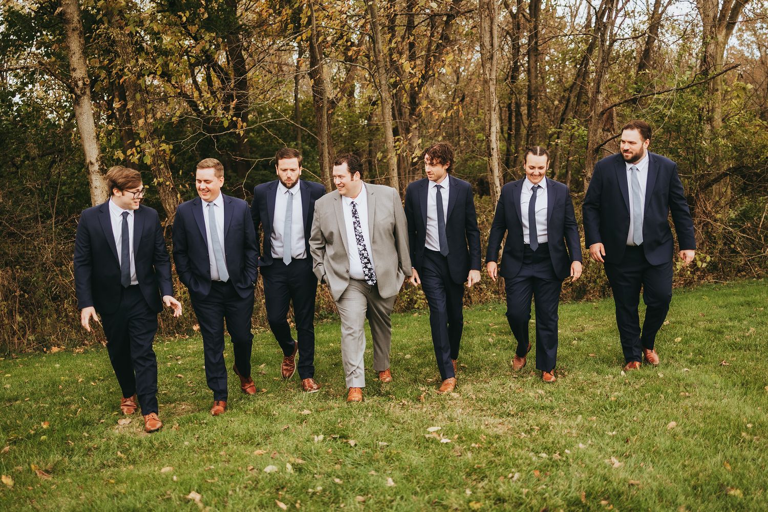Groomsmen walking together through an autumn forest setting wearing navy suits and brown shoes.