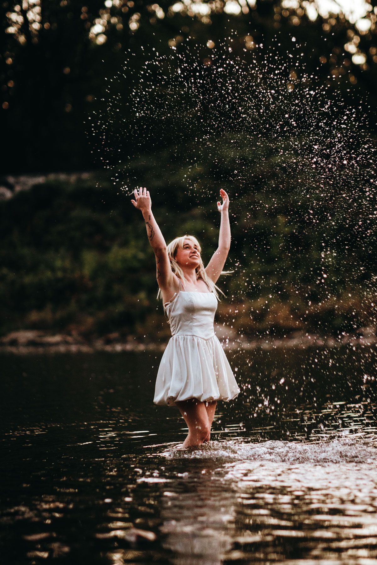 A person in a white dress splashes joyfully in a lake at sunset, creating a sparkling spray of water droplets in the air.