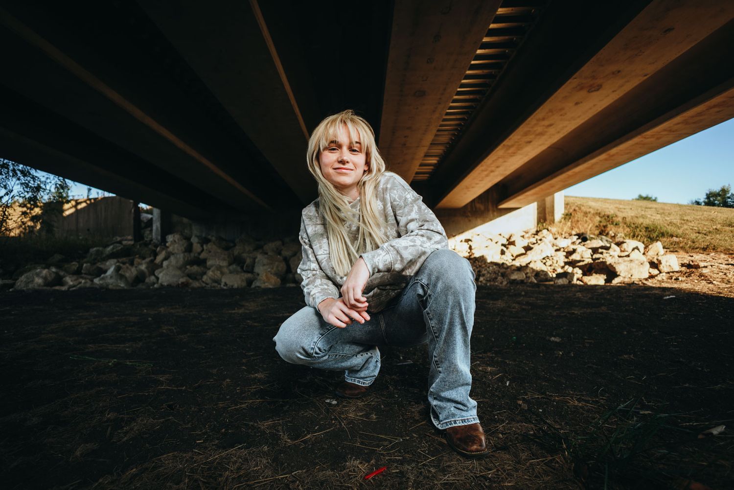 Person in denim clothing sits on rocky ground against a wooden beam structure in natural outdoor setting.