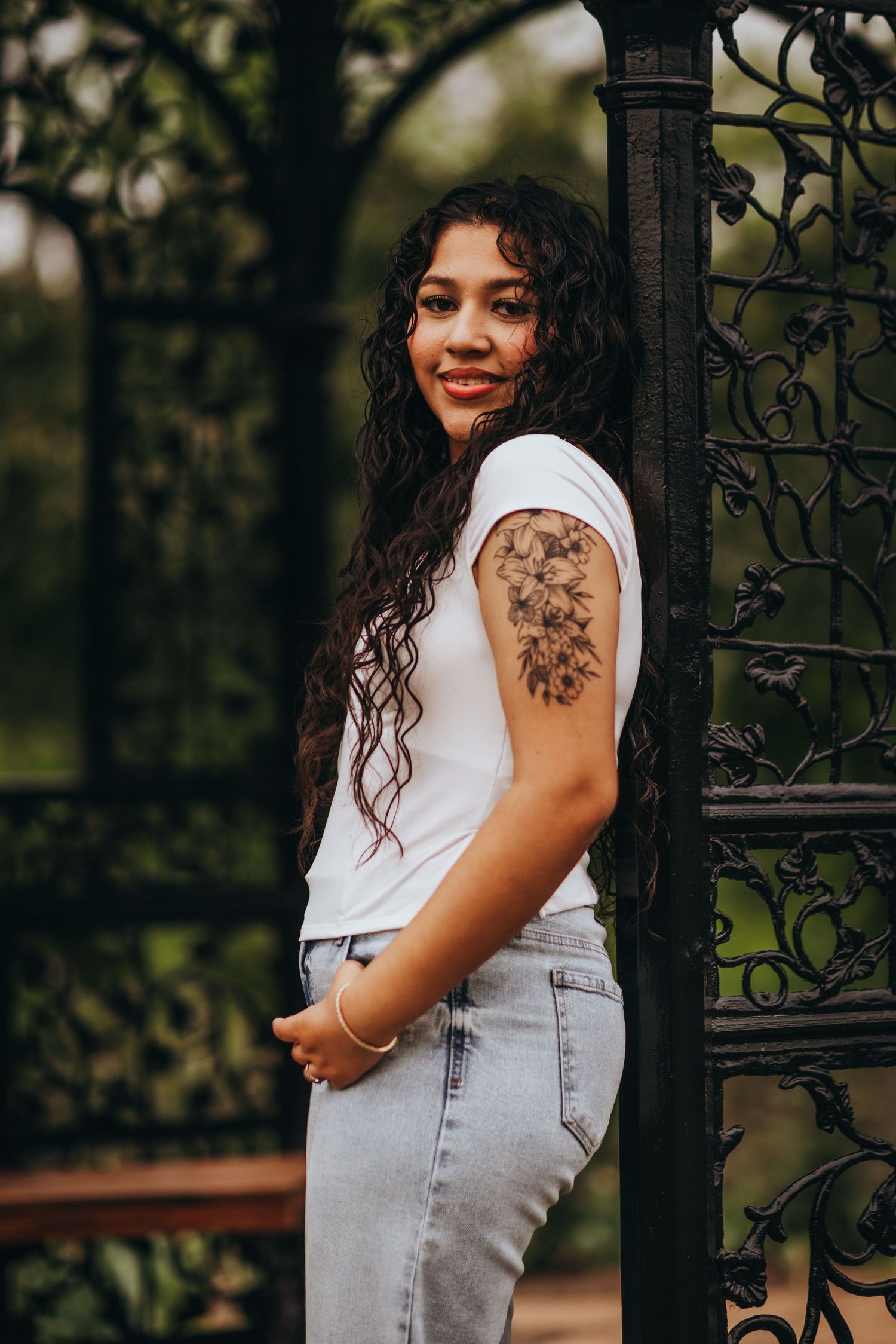 Person with curly hair wearing a white top and light denim overalls poses against an ornate iron gate in a garden setting.