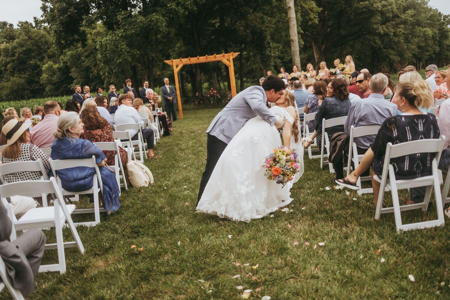 Outdoor wedding ceremony with guests seated on white chairs as couple shares a kiss at the altar.