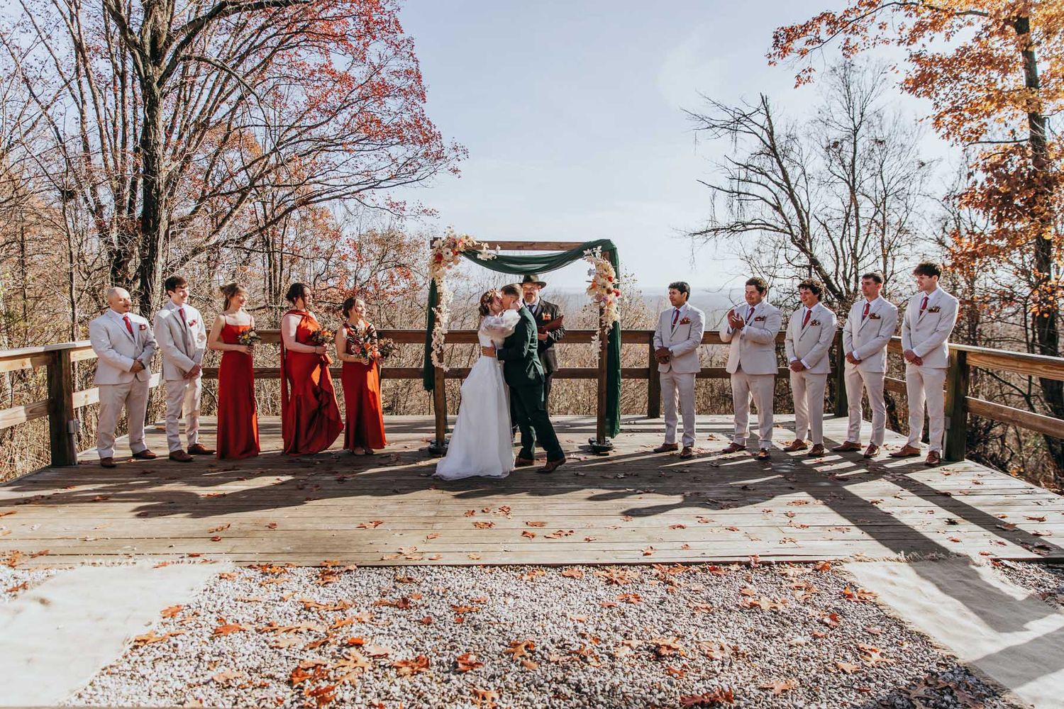 Wedding party members stand in a line outdoors with autumn trees and fallen leaves creating a scenic backdrop.