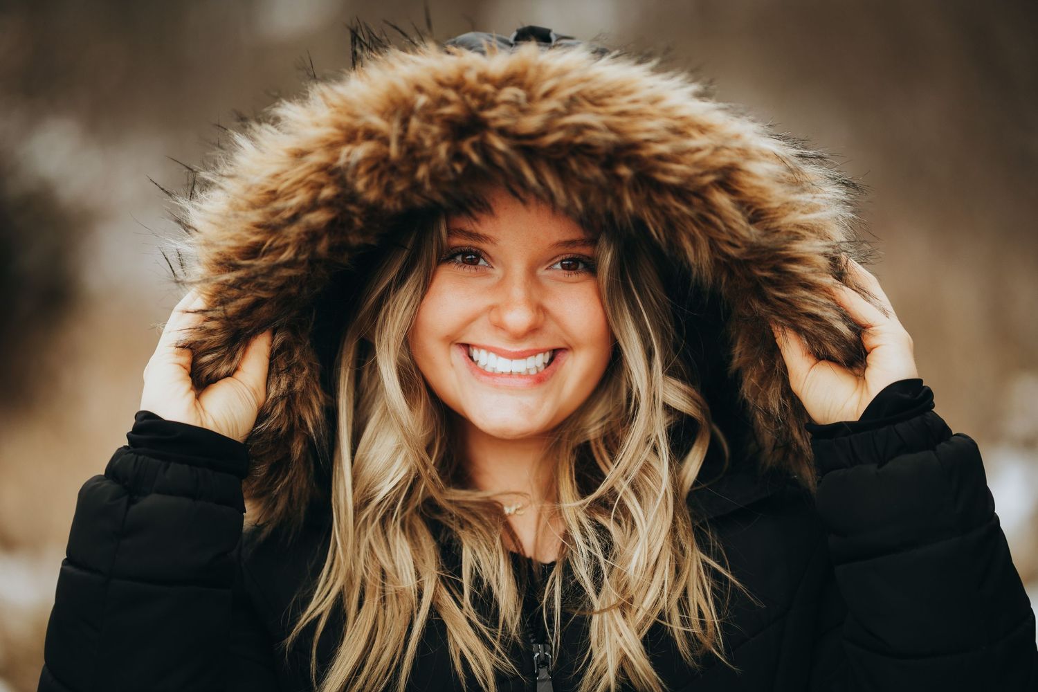 Young woman smiling in a black fur-hooded winter jacket outdoors in a snowy setting.