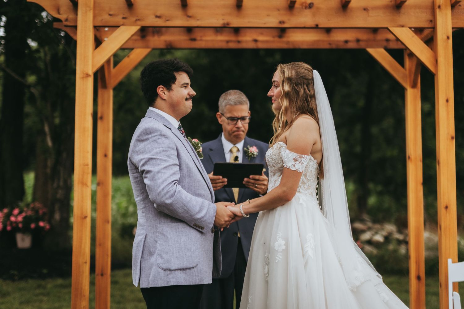Couple exchanges vows under wooden arch at outdoor wedding ceremony with officiant.